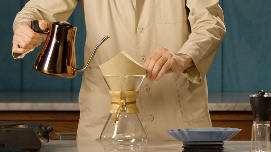 Person making coffee using a pour-over method with a metal kettle and glass carafe.