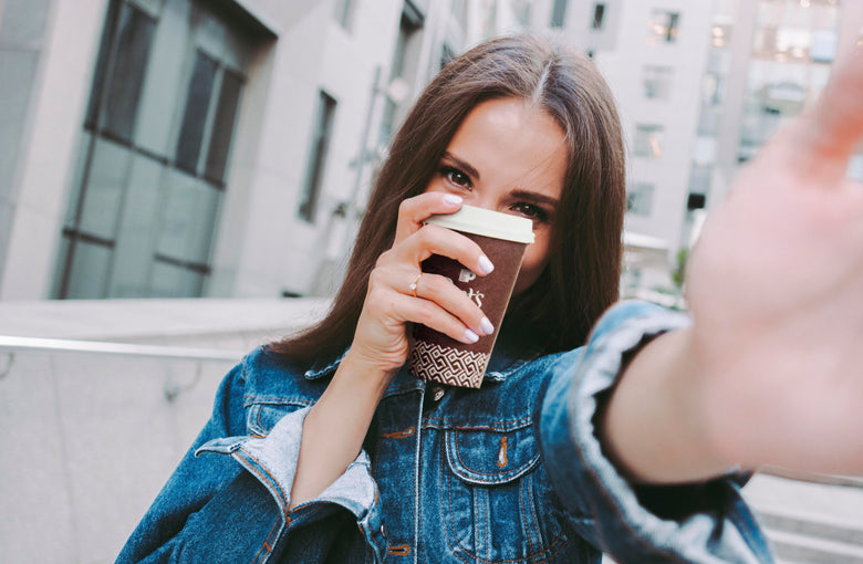 Woman in jean jacket holding Peet's to go cup in front of her face standing in front of a building